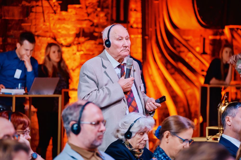 Interior, orange-lit large postindustrial hall. A member of the audience, an older man in a grey suit with headphones on his head, stands up with the microphone, asking the question to the panelists on stage.