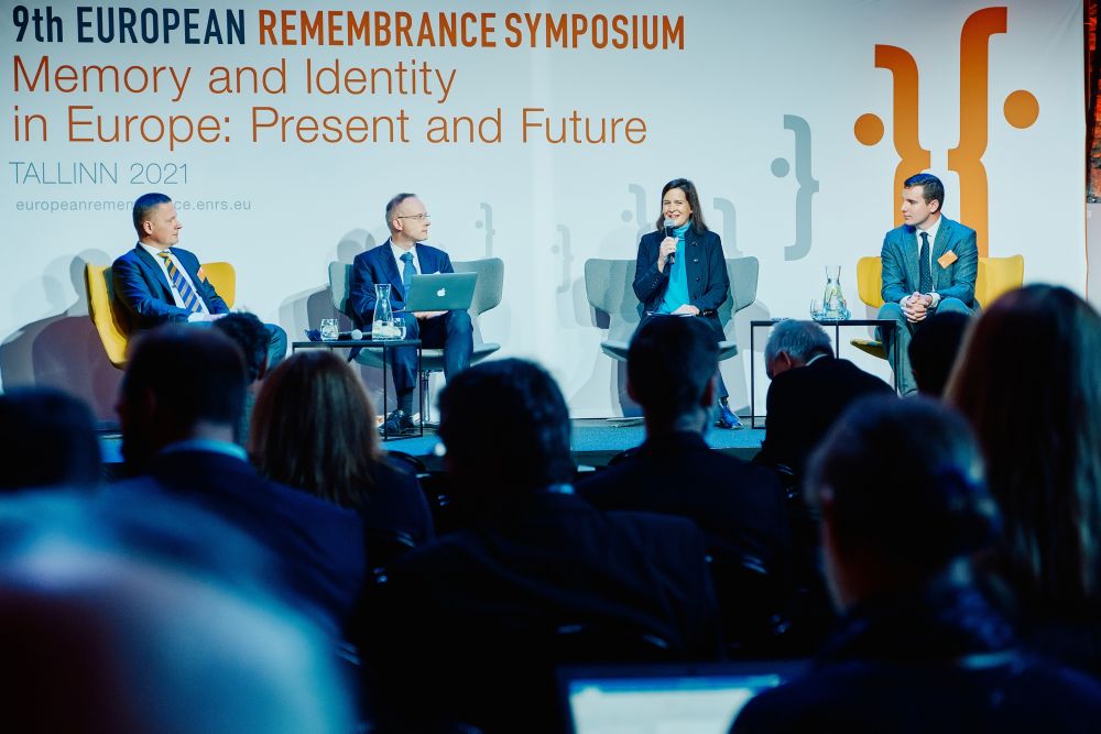 Photo taken from behind the seated auditorium. Rafał Rogulski, Dr Łukasz Kamiński,  Constanze Itzel, and Sergei Metlev sit in the chairs placed on a stage. Behind them, a banner with the Ninth European Remembrance Symposium written on it.
