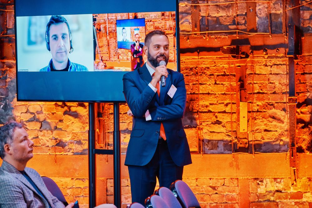 Interior, orange-lit large postindustrial hall. A member of the audience stands up with the microphone, asking the question to the panelists on stage. Behind him, a screen mounted to the wall displays Oriol Lopez Badell and the rest of the audience.