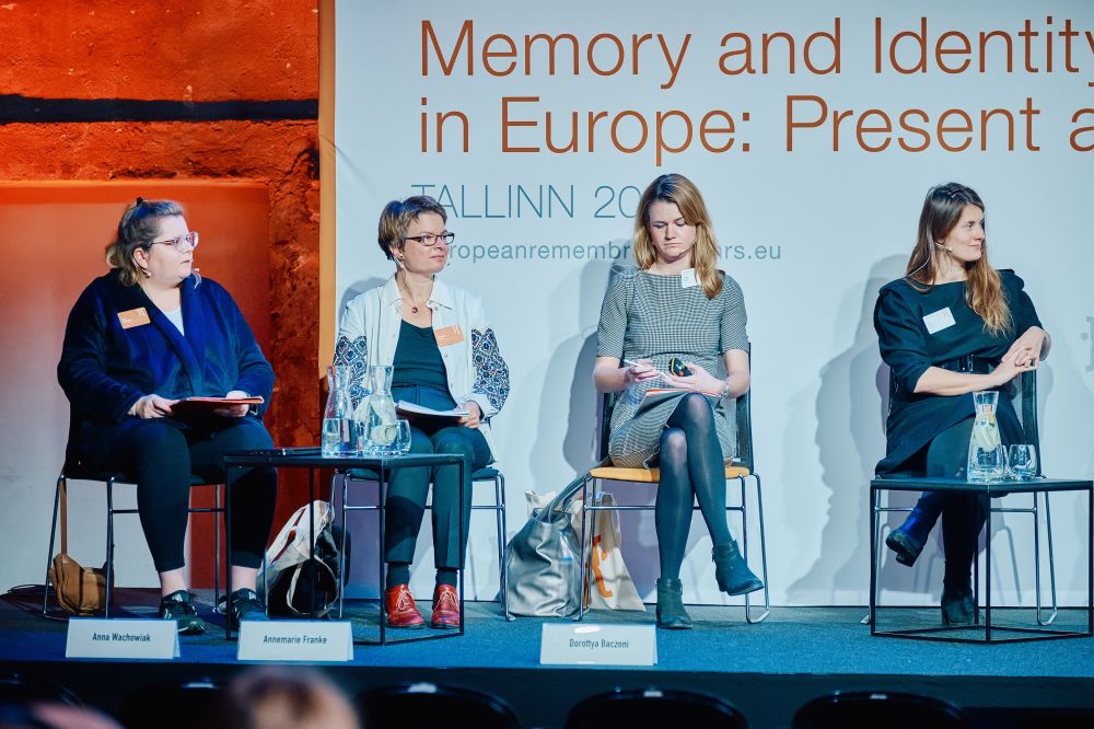 Interior, the main stage of the Ninth European Remembrance Symposium. Four women sit in chairs, with small tables with water and glasses in front of them. Three of them are identified by the small plaques placed in front of them on a stage. From left to right: Anna Wachowiak, Annemarie Franke, and Dorottya Baczoni. On their right, an unidentified woman.