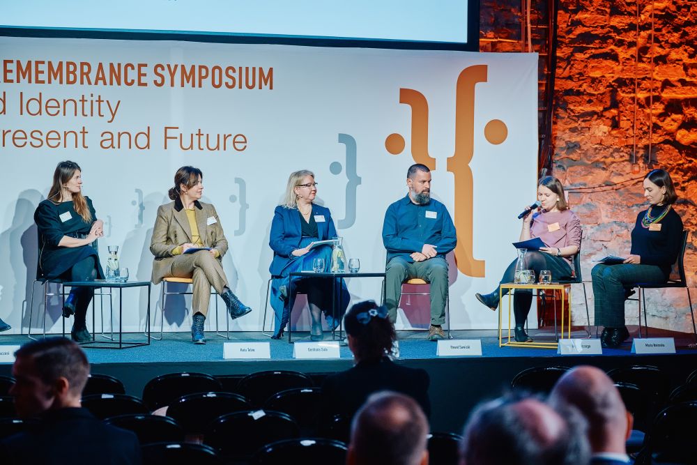 Interior, the main stage of the Ninth European Remembrance Symposium. Six panel participants, five women and one man, sit in chairs with small tables with water and glasses in front of them. The woman who sits on a second to left chai holds a microphone. The rest of the participants listens to her.