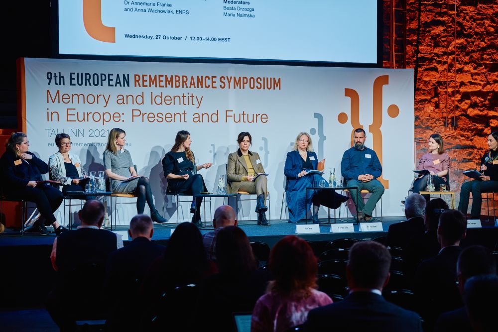 Interior, the main stage of the Ninth European Remembrance Symposium. All nine participants of the panel are sitting in a chair on a stage. They are engaging in a debate. In front of them, the rows of the sited audience.