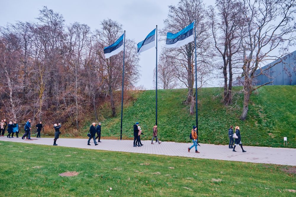 Outdoors, cloudy day. A column of visitors follows a gravel path through the green park. Along their path, leafless trees, and three high poles with Estonian flags.