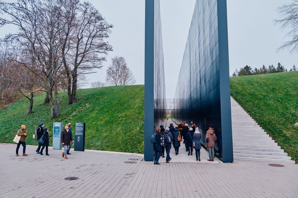 Outdoors, cloudy day. Two black walls divide a green mound, creating a large trench that goes through it. Inside that trench, a group of visitors had gathered. They are following a guide, who faces the camera addressing the crowd.