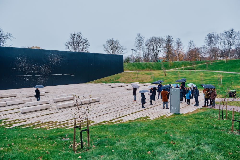 Outdoors, cloudy day. An irregular concrete square with some benches is surrounded by green slopes of small hills. A group of visitors stands around the square. On their left, a large black wall of the architectural installation.