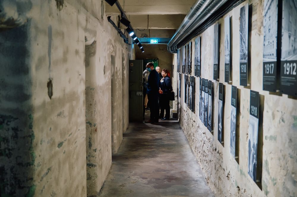 A narrow corridor with bare grey walls. On the left, multiple cell entrances, blocked by the heavy metal doors. On the right wall, black-and-white photographs of the place. Further down the corridor, a group of visitors.