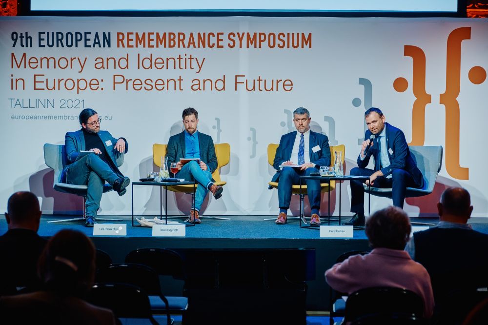 Dr Lars Fredrik Stöcker, Dr Tobias Rupprecht,  Dr Paweł Ukielski, and Dr Peter Jašek sit in the chairs placed on a stage. Behind them, a banner with the Ninth European Remembrance Symposium written on it. In front of them, two small tables with water and glasses.
