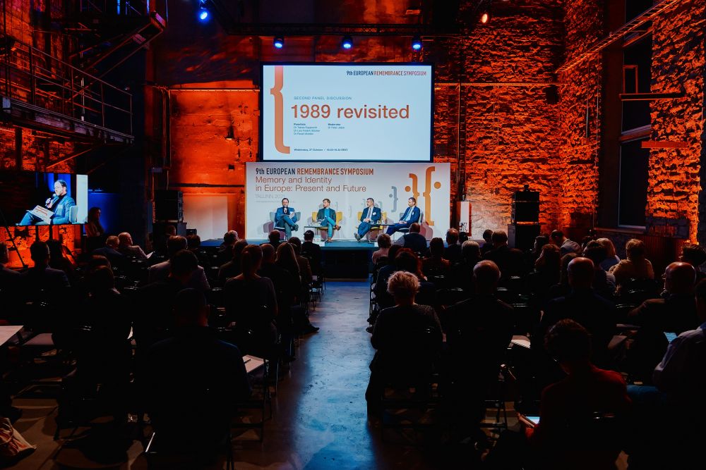 Interior, orange-lit large postindustrial hall. A large audience sits in the dark, listening to the panelists. Further in the back, in the center of the photograph, a fully lit stage of the Ninth European Remembrance Symposium. On it, four men sitting in chairs.