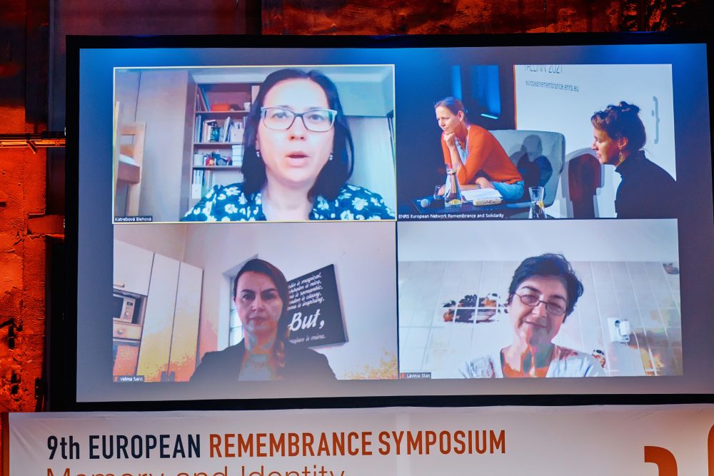 Pull down projector screen above the main stage of the Ninth European Remembrance Symposium. Its display is divided into four, showing Dr Beáta Blehová,  Velma Šarić, and Prof. Lavinia Stan, who are joining the pannel online, as well as the view at the stage in Tallinn, where Prof. Linda Kaljundi and Dr Réka Földváryné Kiss are seating.