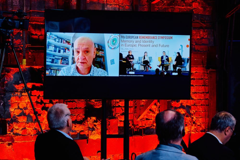 Interior, large postindustrial hall.  A large screen mounted to the orange-lit wall next to the stage. Displayed on it is a split-screen between Dariusz Rosiak, who is joining the panel online, and the stage at the venue of the Ninth European Remembrance Symposium in Tallinn, where the four other panelists are sitting.