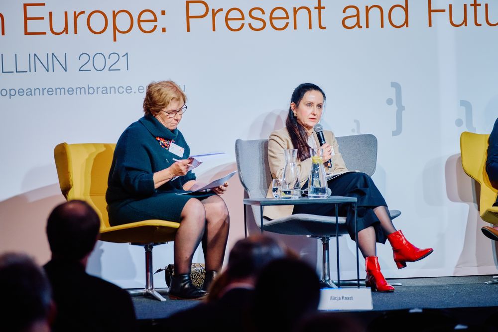 The main stage during a discussion panel. Two women sit on a stage. On a left, Barbara Walshe is looking in concentration at her notes. On a right, Alicja Knast is addressing the audience, holding a microphone. Behind them, a white banner with the Ninth European Remembrance Symposium written on it.
