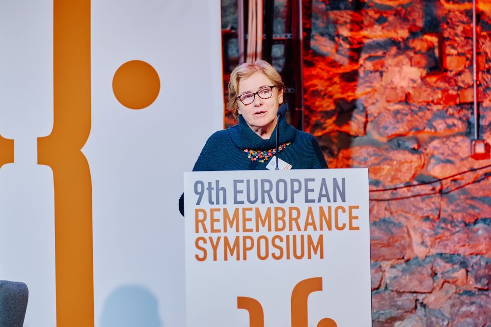 Interior, large postindustrial hall.  Barbara Walshe, Chair of Glencree Centre for Peace and Reconciliation, speaks in front of a packed auditorium. She stands behind the lectern on a spacious stage, with the Ninth European Remembrance Symposium written on a board and an orange-lit brick wall behind her.