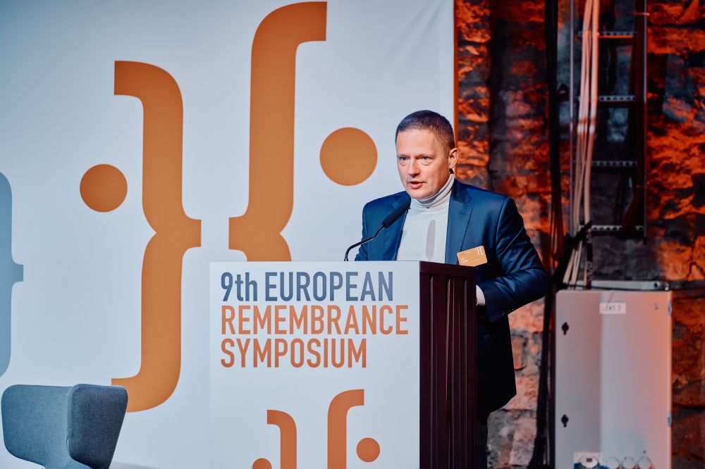 Interior, large postindustrial hall. Rafał Rogulski, ENRS Director, facing the camera speaks in front of a packed auditorium. He stands behind the lectern on a spacious stage, with the Ninth European Remembrance Symposium written on a board behind him.