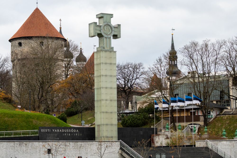 Freedom Square in Tallinn. Grey pillar with the cross on top of it of the War of Independence Victory Column in the foreground. Greenery of Harjumägi park in the background.