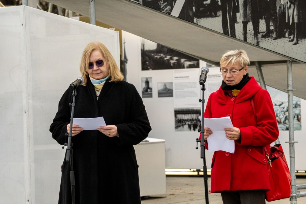 Two middle-aged women, one in a black and the other in a red coat, read into microphones. In the background is the wall of the After the Great War exhibit.