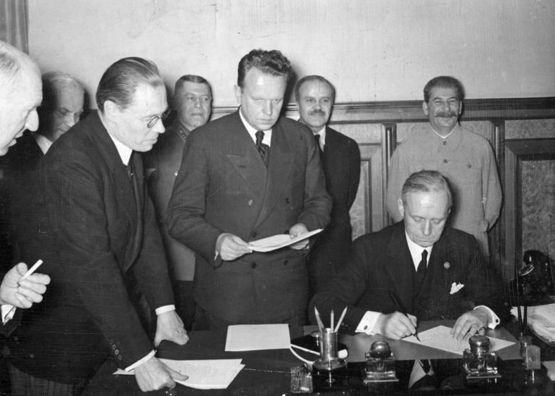 Joachim von Ribbentrop, foreign minister of the Third Reich, signs the Treaty of Non-Aggression between Germany and the Union of Soviet Socialist Republics.  Joseph Stalin and Vyacheslav Molotov in the background. Source: Bundesarchiv, Bild 183-S52480 / CC-BY-SA 3.0